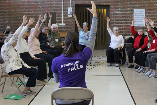 A seated woman leading a group of seated people in arm stretches