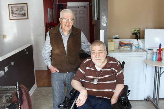 Two smiling older men; one standing and one in a wheelchair