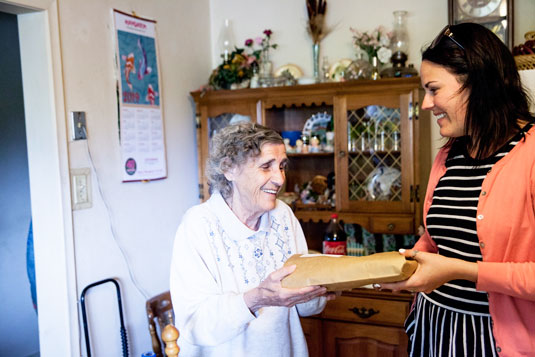 A smiling woman delivering a meal to an older smiling woman in her home