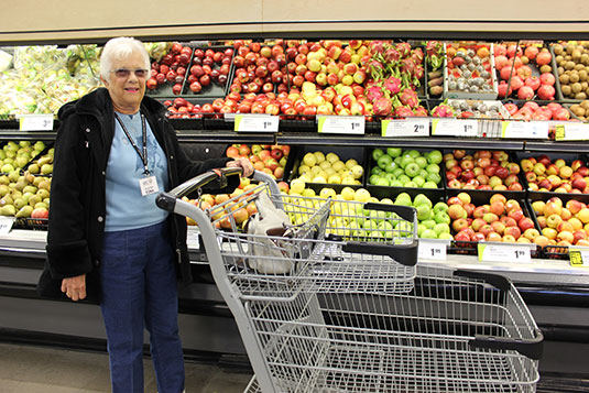 An older woman with a shopping cart in front of a fruit aisle