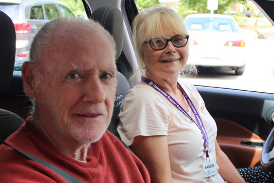 A female volunteer and an older male client in the front seats of a car